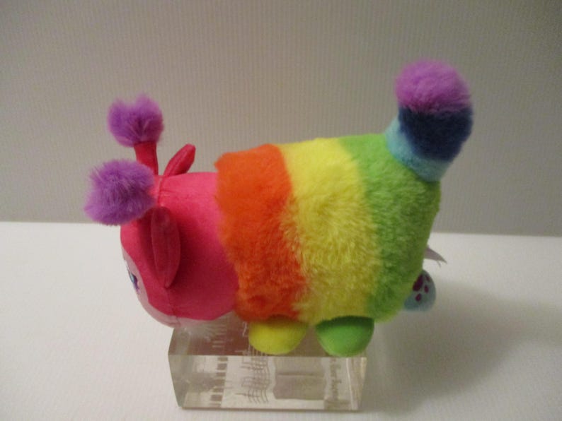 Pu&ograve; includere: Un peluche a forma di bruco con una testa rosa brillante, due antenne a pon-pon viola e un corpo caratterizzato da strisce orizzontali arancioni, gialle, verdi e blu. La coda &egrave; un ciuffo soffice viola e blu, ed &egrave; dotato di piccoli piedini.