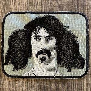 Puede incluir: Parche bordado con un retrato de un hombre con pelo largo y oscuro y un bigote prominente. El parche tiene un fondo azul claro y un borde negro. El retrato está realizado en hilo blanco y negro.