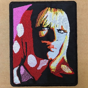 May include: Embroidered patch featuring a stylized portrait of a person with blonde and orange hair, and blue eyes. The design includes a red and white polka-dotted element on the left side, set against a black background. The patch is rectangular.