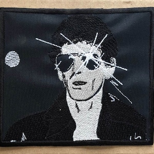 May include: Black and white embroidered patch of a man wearing sunglasses with a white background behind him. The man's eyes are covered by the sunglasses and there are white lines radiating from his eyes.