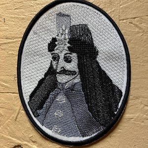 Può includere: Toppa ricamata di forma ovale con il ritratto di un uomo con grandi baffi, che indossa un cappello e lunghi capelli neri. Il ritratto è nei toni del grigio, nero e blu, con un bordo nero.