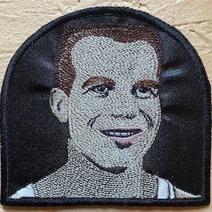 May include: Embroidered patch featuring a portrait of a smiling person with brown hair and a white shirt. The patch has a black border and a semi-circular top. The portrait is detailed with various thread colors.