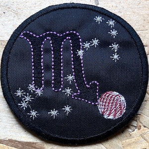Op de afbeelding: Een zwarte geborduurde patch met een paarse omtrek van het sterrenbeeld Schorpioen, met een rode geborduurde cirkel die een planeet voorstelt. De achtergrond is zwart met witte geborduurde sterren.