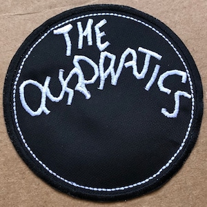 May include: A black and white embroidered patch with the text "THE QUADRATS" in a circular shape.