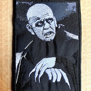 Könnte beinhalten: Gestickter Aufnäher mit einem Schwarzweißbild eines Vampirs mit scharfen Zähnen und Krallen. Der Vampir trägt einen dunklen Anzug und hat einen blassen Teint.