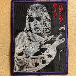 May include: Black embroidered patch with a white image of a person playing a guitar. The text "RICK " and "WAKENSS" is embroidered in red on the right side of the patch.