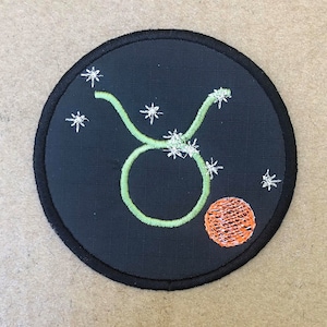 Op de afbeelding: Een zwarte geborduurde patch met een groen Stier-sterrenbeeld en een kleine oranje cirkel. De patch is omgeven door een zwarte rand.