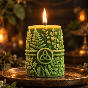 Könnte beinhalten: Eine zylindrische grüne Kerze mit brennendem Docht, verziert mit geprägten Farn- und Wildblumenmustern. Ein kreisförmiges Triquetra-Symbol ist mittig auf der Vorderseite platziert und von einem seilartigen Band umgeben. Die Kerze steht auf einem Metallteller.