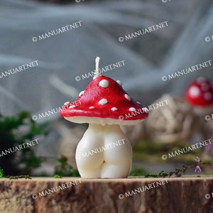 May include: A white and red mushroom-shaped candle with white dots on the top. The candle is sitting on a wooden surface with green moss and a small brown pine cone.