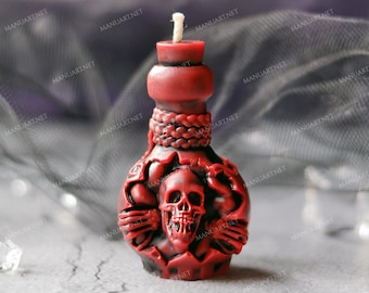Pode incluir: Uma vela vermelha em forma de garrafa com um design de caveira. A vela tem um pavio branco e está sobre uma superfície cinza.
