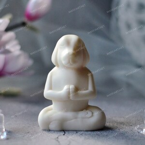 May include: A white dog figurine in a seated yoga pose with hands together in prayer. The figurine is on a gray surface with a blurred background of flowers and decorative elements. Clear, ice-like crystals are in the foreground.