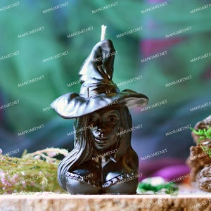 Peut inclure: Bougie noire en forme de sorcière avec un chapeau pointu et de longs cheveux. La bougie est posée sur une surface en bois avec de la végétation verte en arrière-plan.