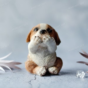 Può includere: Una piccola figurina in ceramica di un cucciolo seduto, di colore marrone e bianco, con le zampe che gli coprono la bocca. La figurina è su uno sfondo blu chiaro.