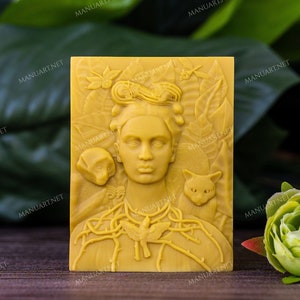 Peut inclure: Une sculpture en cire d'abeille jaune de Frida Kahlo avec un chat et un crâne. La sculpture est sur une surface en bois avec des feuilles vertes en arrière-plan.