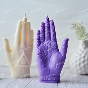 May include: Two hand-shaped candles, one purple and one white. The purple candle has astrological symbols and lines on it. The white candle has a triangle with an eye inside. Both candles are on a wooden surface.