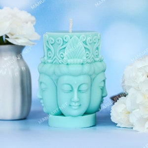 Può includere: Una candela blu turchese con tre volti di Buddha scolpiti sul davanti. La candela è appoggiata su una superficie blu chiaro con garofani bianchi in un vaso bianco.