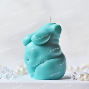 Könnte beinhalten: Eine tealblaue Kerze in Form eines menschlichen Körpers. Die Kerze steht auf einer weißen Oberfläche mit weißen Perlen und klaren Glassteinen drumherum.