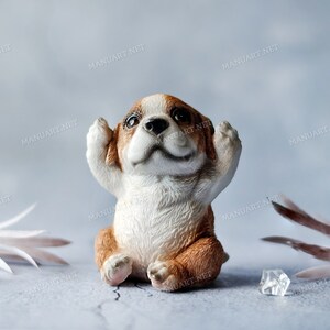 Può includere: Statuetta di cane in ceramica in posizione seduta, con le zampe alzate verso le orecchie. Il cane ha pelo marrone e bianco, con un'espressione carina. Lo sfondo è azzurro chiaro, con elementi decorativi e cubetti di ghiaccio.