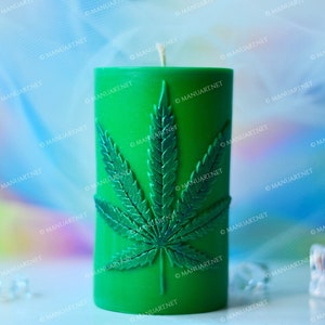 Può includere: Candela a pilastro verde con un designo in rilievo dettagliato di una foglia di marijuana. La candela è su una superficie bianca con uno sfondo sfocato di colori iridescenti.