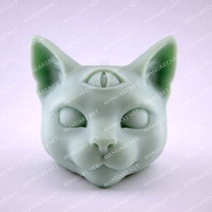 May include: A light green ceramic cat head sculpture with three eyes. The eyes are black and white and the cat has a small, pointed nose.