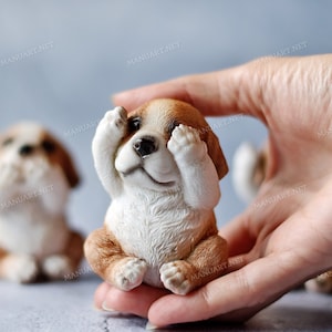 Può includere: Una piccola statuetta di ceramica bianca e marrone di un cane che si copre gli occhi con le zampe. La statuetta è tenuta nella mano di una persona.