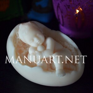 May include: A white baby figurine sleeping inside a cracked egg shell. The baby is holding a small object in its hand. The figurine is made of a smooth, white material.