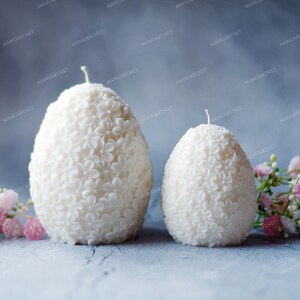 May include: Two white egg-shaped candles with a floral design. The candles are sitting on a gray surface with pink flowers.