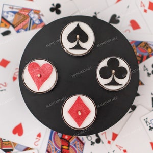 Puede incluir: Un portavelas circular negro con cuatro velas, cada una con un símbolo de palo de la baraja: picas, corazones, tréboles y diamantes. El portavelas está rodeado de naipes.