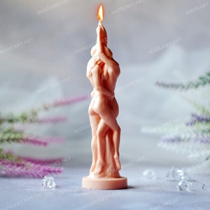 May include: A light peach-coloured pillar candle shaped like two figures embracing in a standing position. The candle features a smooth, sculpted wax surface with detailed human forms and is lit at the top, producing a small, bright orange flame.