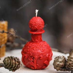 Può includere: Candela rossa a forma di bottiglia con intricati disegni floreali e la parola "Pozione" incisa sul davanti. La candela è su una superficie di legno con pigne.
