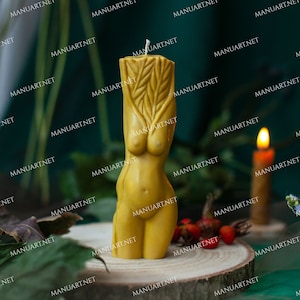 May include: A yellow beeswax candle shaped like a human figure with a detailed design on the torso. The candle is sitting on a wooden slice with green foliage and red berries in the background.
