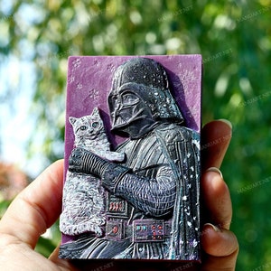 Peut inclure: Un savon violet et noir avec une image en relief de Dark Vador tenant un chat. Le chat est blanc et gris avec des yeux bleus.