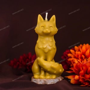 Könnte beinhalten: Eine gelbe Bienenwachskerze in Form eines Fuchses, der auf einem Holzsockel sitzt. Der Fuchs hat einen Ausdruck mit geschlossenen Augen und einen eingerollten Schwanz. Die Kerze ist von orangefarbenen und roten Blumen umgeben.