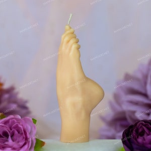 Peut inclure: Une bougie beige en forme de pied humain. La bougie est debout sur une surface blanche avec des fleurs violettes et orange en arrière-plan.