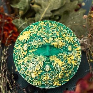 Könnte beinhalten: Runde, grün-goldene dekorative Plakette mit einem Green Man-Gesicht. Das Gesicht ist von Eichenblättern und Eicheln umgeben. Die Plakette befindet sich vor einem Hintergrund aus getrockneten Blumen und Blättern.