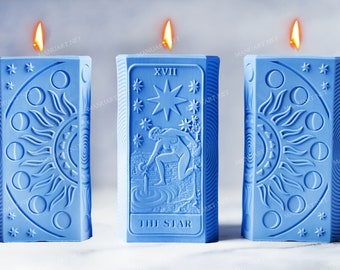 Puede incluir: Tres velas azules con diseños de cartas de tarot. La vela del medio presenta la carta de la Estrella con el número romano XVII y el texto "The Star". Las velas están encendidas.