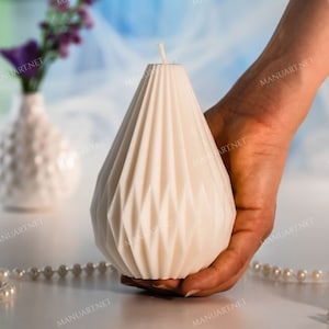 Puede incluir: Una vela blanca con forma de lágrima y un patrón geométrico. La vela está siendo sostenida por una mano, y hay perlas blancas y una flor morada sobre una superficie blanca.