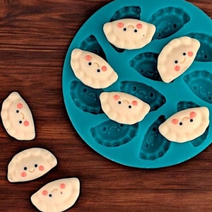 Könnte beinhalten: Eine blaue Silikonform mit acht einzelnen Vertiefungen in Form von Halbmonden. Die Form ist mit weißem Teig gefüllt, der die Form von Halbmonden mit rosa lächelnden Gesichtern hat. Auf einer Holzoberfläche liegen außerdem sieben weitere weiße Teig-Halbmonde mit rosa lächelnden Gesichtern.