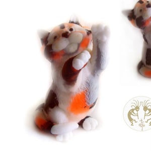Pode incluir: Uma figurinha de cerâmica de um gato calico com marcas laranja, pretas e brancas. O gato está em pé sobre suas patas traseiras com suas patas dianteiras levantadas no ar. A figurinha é uma representação caprichosa e brincalhona de um gato.