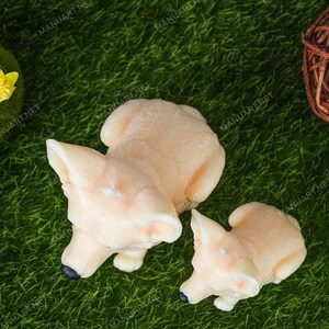 May include: Two ceramic fox figurines, one larger than the other, are sitting on a green artificial grass surface. The larger fox is light peach in color and the smaller fox is a slightly darker peach color. Both foxes have black eyes and noses.