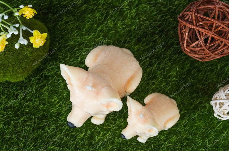 May include: Two ceramic fox figurines, one larger than the other, are sitting on a green artificial grass surface. The larger fox is light peach in color and the smaller fox is a slightly darker peach color. Both foxes have black eyes and noses.