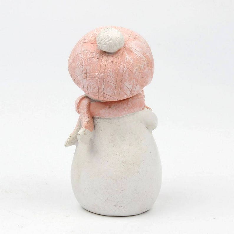 May include: A small white snowman figurine wearing a pink knit hat with a white pom pom and a pink scarf.