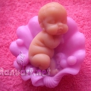 Puede incluir: Una pequeña figurita de bebé durmiendo hecha de jabón rosa, anidada en una concha rosa con detalles de perlas.
