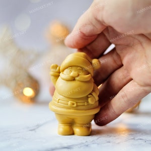 May include: A golden yellow Santa Claus figurine with a white beard and a red hat. The figurine is holding a bag in one hand and is standing on a white surface.