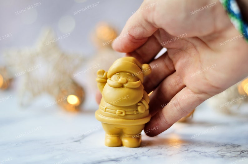 May include: A golden yellow Santa Claus figurine with a white beard and a red hat. The figurine is holding a bag in one hand and is standing on a white surface.