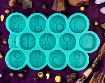 Könnte beinhalten: Tealfarbene Silikonform mit 12 runden Vertiefungen, jede mit einem anderen astrologischen Symbol. Die Form befindet sich auf einer violetten Oberfläche mit verschiedenen Dekorationsartikeln.