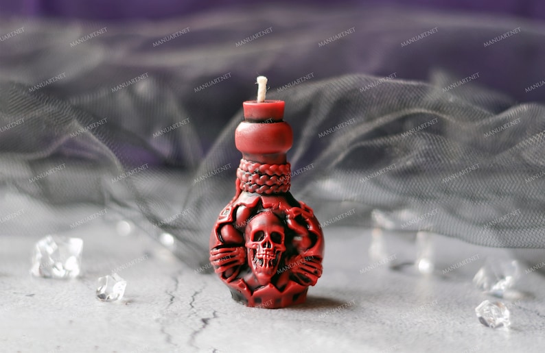 Pode incluir: Uma vela vermelha em forma de garrafa com um design de caveira. A vela tem um pavio branco e est&aacute; sobre uma superf&iacute;cie cinza.