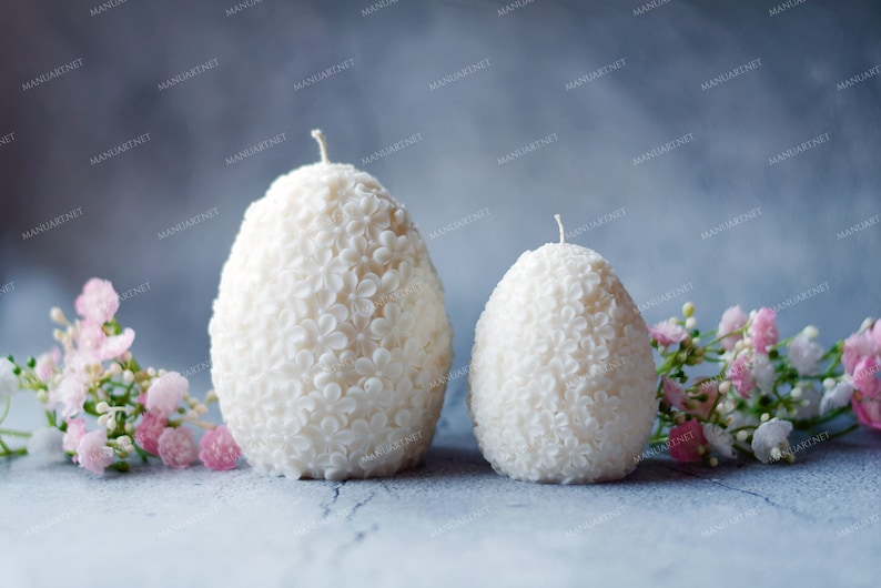 May include: Two white egg-shaped candles with a floral design. The candles are sitting on a gray surface with pink flowers.