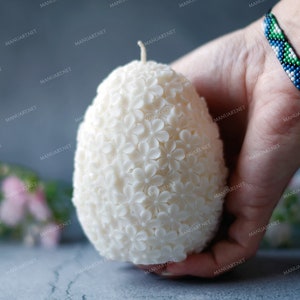 May include: A white egg-shaped candle with a floral design. The candle is made of wax and has a small wick on top. The candle is being held in a hand.