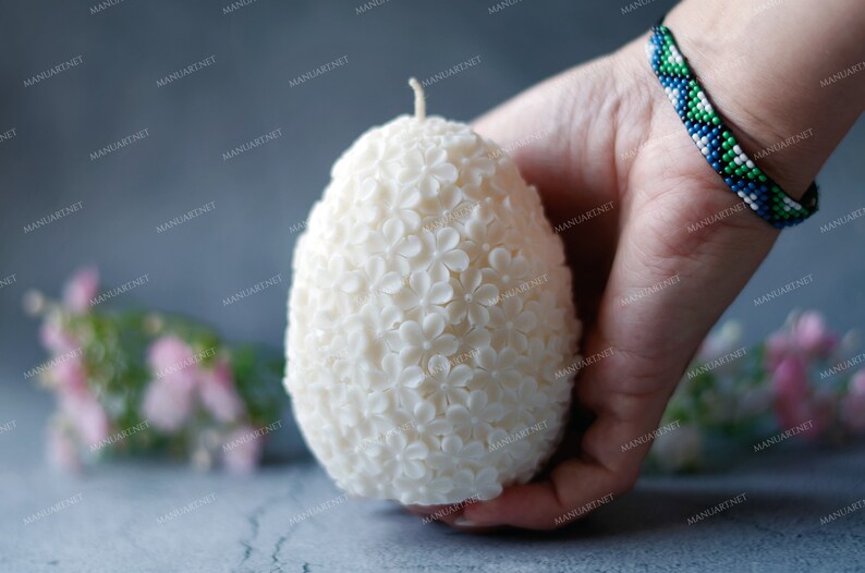 May include: A white egg-shaped candle with a floral design. The candle is made of wax and has a small wick on top. The candle is being held in a hand.
